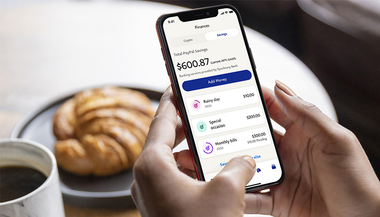 إنشاء حساب باي بال؛ دليل شامل لمنصة PayPal الرائعة 5 ممبزات ابلكيشن باي بال وكيفية التسجيل وتظهر يد شخص تمسك بهاتف محمول عليه التطبيق ويتناول وجبة الافطار